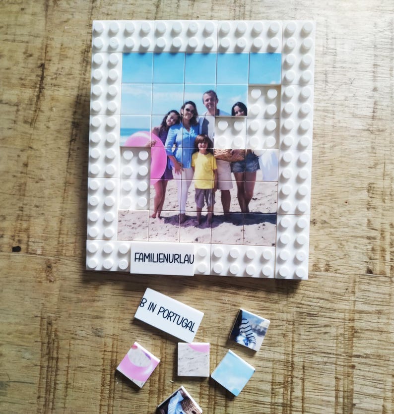 May include: A white plastic building block frame containing a photo mosaic of a family on a beach. The frame features a white tile with the text FAMILIENURLAU. Several loose square tiles with photo fragments and the text B IN PORTUGAL are below.