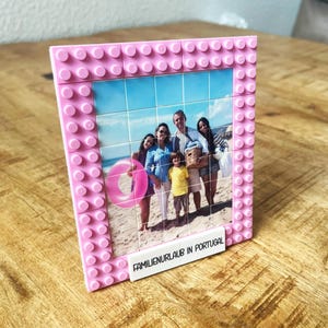 May include: A rectangular picture frame constructed from pink plastic building blocks. The frame holds a grid of square photo tiles depicting a family on a beach with a pink inner tube. A white block at the bottom displays the text FAMILIENURLAUB IN PORTUGAL.