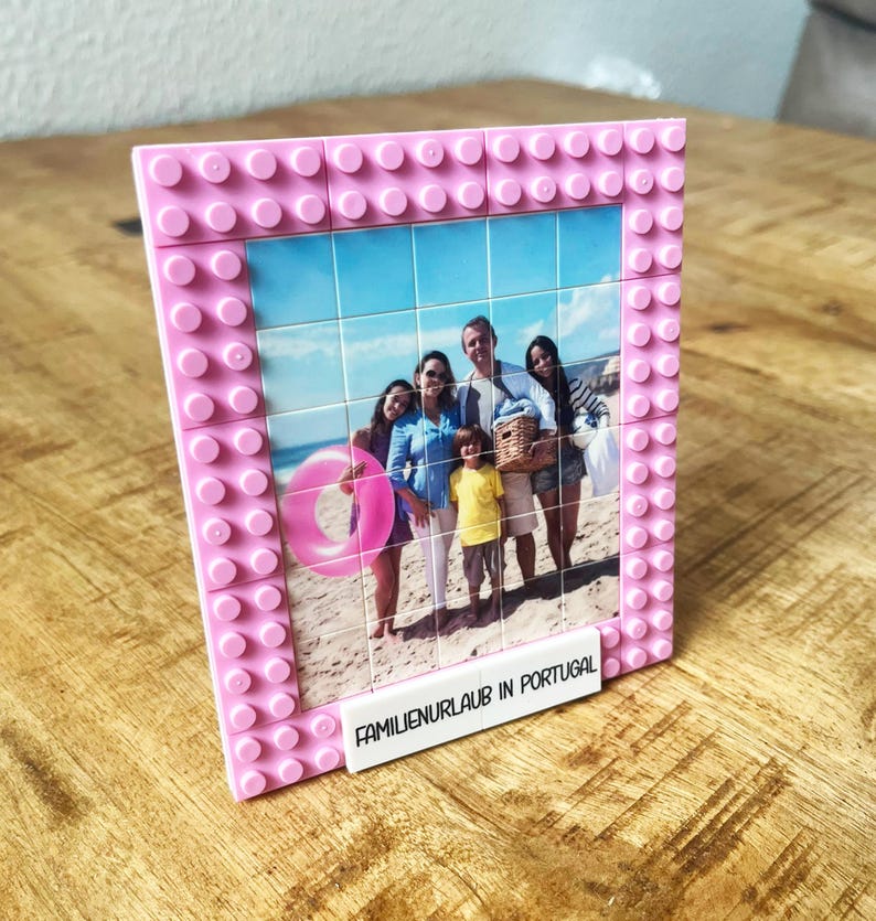 May include: A rectangular picture frame constructed from pink plastic building blocks. The frame holds a grid of square photo tiles depicting a family on a beach with a pink inner tube. A white block at the bottom displays the text FAMILIENURLAUB IN PORTUGAL.