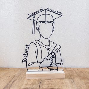 May include: A black line art graduation figure with a mortarboard hat and diploma, mounted on a wooden base. The text "Master of Science 2025" is above the figure's head, and the name "Robert" is on the side.