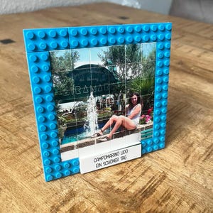 May include: A square picture frame constructed from bright blue interlocking plastic building bricks. The frame holds a grid of printed tiles displaying a photograph of a person sitting by a fountain. A white rectangular tile at the base reads CAMPOMARINO LIDO EIN SCH&Ouml;NER TAG.