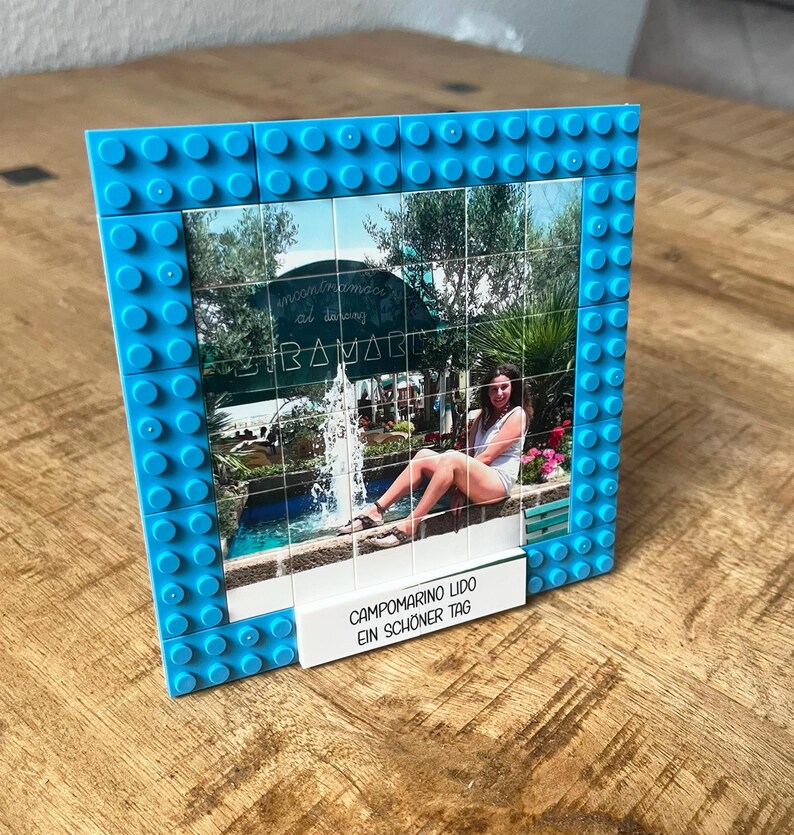 May include: A square picture frame constructed from bright blue interlocking plastic building bricks. The frame holds a grid of printed tiles displaying a photograph of a person sitting by a fountain. A white rectangular tile at the base reads CAMPOMARINO LIDO EIN SCH&Ouml;NER TAG.