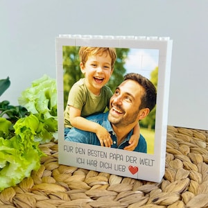 May include: White block photo frame featuring a smiling man and child. The frame has the text "F&Uuml;R DEN BESTEN PAPA DER WELT ICH HAB DICH LIEB" with a red heart. The background is a natural woven mat.