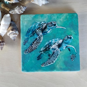 May include: Two blue sea turtles swimming in a turquoise blue ocean. The turtles are painted on a square, light blue stone coaster.