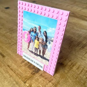 May include: A pink picture frame constructed from interlocking plastic building blocks. The frame holds a grid-like photograph of four people on a beach with a pink inner tube. A white rectangular tile at the bottom displays the text FAMILIENURLAUB IN PORTUGAL.