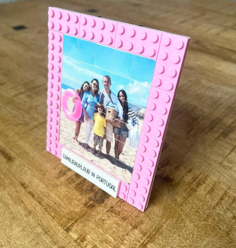 May include: A pink picture frame constructed from interlocking plastic building blocks. The frame holds a grid-like photograph of four people on a beach with a pink inner tube. A white rectangular tile at the bottom displays the text FAMILIENURLAUB IN PORTUGAL.