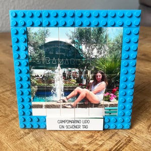 May include: A square photo frame constructed from bright blue interlocking plastic building bricks. The frame displays a printed image of a person sitting by a fountain, with a white base plate featuring the text CAMPOMARINO LIDO EIN SCH&Ouml;NER TAG.