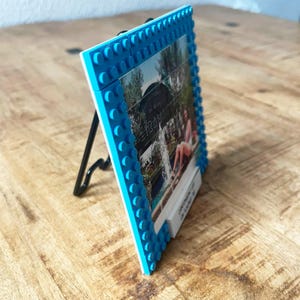 May include: A square picture frame constructed from bright blue interlocking plastic building bricks. The frame features a raised stud texture along the border and is supported by a thin black metal easel stand. A photograph is displayed inside.
