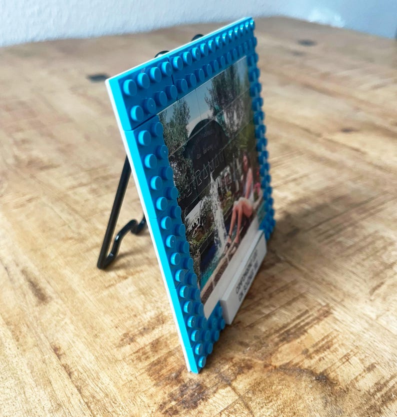 May include: A square picture frame constructed from bright blue interlocking plastic building bricks. The frame features a raised stud texture along the border and is supported by a thin black metal easel stand. A photograph is displayed inside.