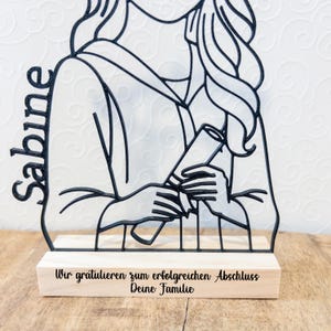 May include: A black metal graduation figure with the name "Sabine" on the left side. The figure holds a diploma and is mounted on a wooden base with the text "Wir gratulieren zum erfolgreichen Abschluss Deine Familie".