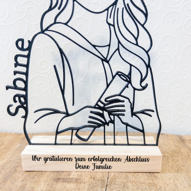 May include: A black metal graduation figure with the name "Sabine" on the left side. The figure holds a diploma and is mounted on a wooden base with the text "Wir gratulieren zum erfolgreichen Abschluss Deine Familie".