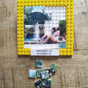 May include: A square yellow plastic building block frame containing a photo mosaic of a person sitting by a fountain. A white rectangular tile at the bottom displays the text CAMPOMARINO LIDO EIN SCH&Ouml;NER TAG. Several loose photo-printed tiles lie below.