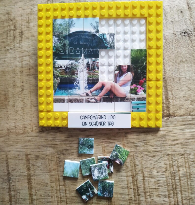 May include: A square yellow plastic building block frame containing a photo mosaic of a person sitting by a fountain. A white rectangular tile at the bottom displays the text CAMPOMARINO LIDO EIN SCH&Ouml;NER TAG. Several loose photo-printed tiles lie below.