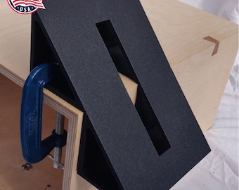 May include: A black woodworking jig with a rectangular opening, used to create a 90-degree angle. The jig is clamped to a piece of wood.