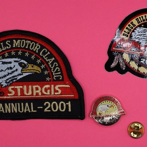 Peut inclure: Trois patchs du rassemblement motocycliste de Sturgis. L'un est un patch brodé noir et rouge avec un aigle chauve et le texte "Black Hills Motor Classic" et "Sturgis 61st Annual - 2001". Le second est un autocollant noir et rouge avec un aigle chauve et le texte "Black Hills Motor Classic" et "Sturgis 2001". Le troisième est une épingle en métal doré avec un aigle chauve et le texte "Sturgis 2001".