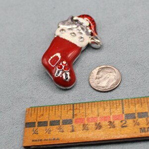 Puede incluir: Un pin de Navidad de color rojo y blanco con forma de calcetín, con pedrería y un gorro de Papá Noel. El pin mide aproximadamente 2,5 cm de alto y se encuentra junto a una moneda de diez céntimos de euro para comparar el tamaño.