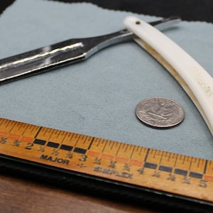 May include: A white handled straight razor with a silver blade. The razor is laying on a blue cloth with a wooden ruler and a US quarter.