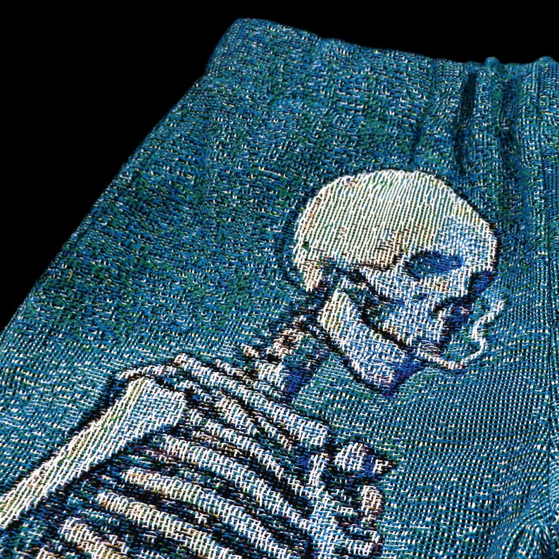 May include: A blue tapestry featuring a skeleton with a cigarette in its mouth. The skeleton is woven into the fabric and has a detailed design.