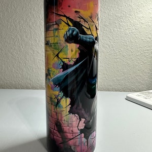 May include: A black and white stainless steel tumbler with a colorful graphic of a superhero breaking through a brick wall. The graphic features shades of pink, yellow, blue, and green.