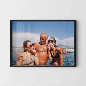 May include: A framed photograph shows three individuals standing in the ocean. A shirtless person with a white beaded cross necklace stands between two others wearing bikini tops and sunglasses. All three are holding cigarettes.