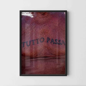 May include: A framed wall art print featuring a close-up of textured, reddish-brown skin with the black text TUTTO PASSA tattooed across the chest area. The artwork is enclosed in a simple black frame, highlighting the contrast between the dark lettering and the skin's natural, weathered appearance.