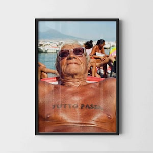 May include: A framed photograph featuring a sun-tanned person wearing dark-rimmed sunglasses. The person has the words "TUTTO PASSA" tattooed across their chest. The background shows a bright, sunny beach scene with water and other people.