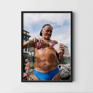 May include: A framed photograph featuring a person wearing a dark baseball cap and blue swim briefs, pouring a bottle of Peroni beer into a white plastic cup while holding a cigarette. The person wears a silver cross necklace.