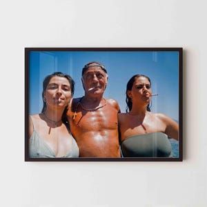 May include: A framed photograph shows three people standing together against a clear blue sky. The person on the left wears a silver top, the center person is shirtless with a beaded necklace, and the person on the right wears a green top.