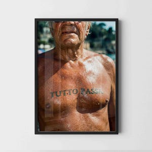 May include: A framed photographic print featuring a sun-tanned torso with a dark tattoo across the chest that reads TUTTO PASSA. The image captures the upper body and neck of an individual with visible skin texture and natural lighting.