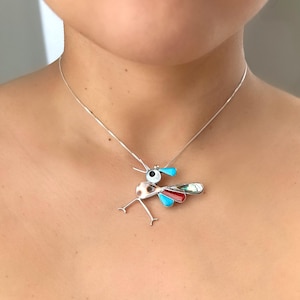 May include: A silver chain necklace with a colorful bird pendant made of turquoise, red, and white shell. The bird has a large eye and a long beak.
