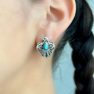 May include: Silver eagle earrings with turquoise stones. The earrings are in the shape of an eagle with its wings spread out. The turquoise stones are set in the center of the eagle's chest.