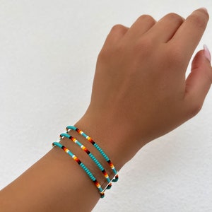 May include: Three beaded bracelets in turquoise, red, yellow, black, and white are stacked on a wrist. The bracelets are made of small, colorful beads strung together. The background is a plain white surface.