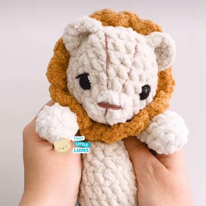 May include: A handmade crochet lion plush toy. The lion has a cream-colored face and body, a brown mane, and black eyes. The text "TWO LITTLE LIONS" is visible on the lower left. The toy is held in two hands.