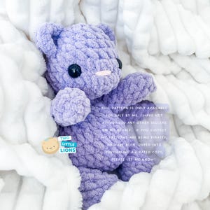 May include: A purple crocheted teddy bear sitting on a white fluffy blanket. The bear has black eyes and a pink nose. The text "TWO LITTLE LIONS" is on a blue and yellow sticker on the blanket. The text "THIS PATTERN IS ONLY AVAILABLE FOR SALE BY ME. I HAVE NOT AUTHORIZED ANY OTHER SELLERS ON MY BEHALF. IF YOU SUSPECT MY PATTERNS ARE BEING PIRATED, OR HAVE BEEN DUPED INTO PURCHASING A PIRATED COPY, PLEASE LET ME KNOW!" is printed on the blanket.