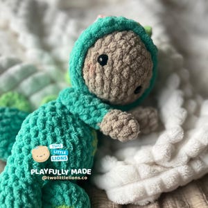 May include: A crocheted green and brown dinosaur plush toy with a pink flower on its head. The toy is lying on a white blanket with the text "TWO LITTLE LIONS PLAYFULLY MADE @twoliltlelions.co" on the side.