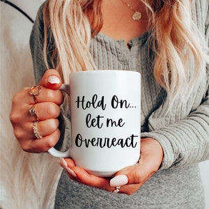 Puede incluir: Taza de café de cerámica blanca con texto negro que dice "Hold On... let me overreact".