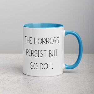 May include: White ceramic coffee mug with a blue rim and handle. The mug has black text that reads "The Horrors Persist But So Do I."