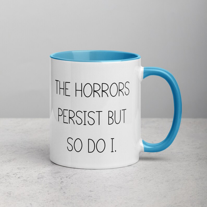 May include: White ceramic coffee mug with a blue rim and handle. The mug has black text that reads "The Horrors Persist But So Do I."