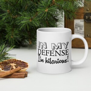 May include: White ceramic mug with black text that reads "In my defense I'm hilarious!"