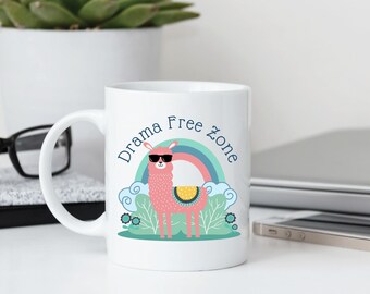 May include: White ceramic mug with a colorful illustration of a pink llama wearing sunglasses in front of a rainbow. The mug has the text "Drama Free Zone" in blue. The mug is on a white surface.