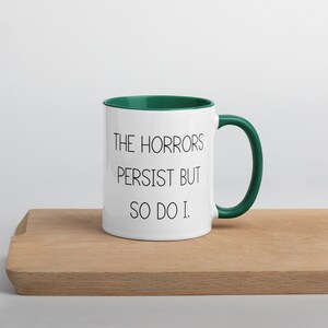 May include: White ceramic coffee mug with a green rim and handle. The mug has black text that reads "The horrors persist but so do I."