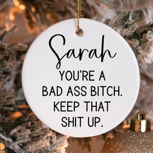 May include: A white ceramic Christmas ornament with the name "Sarah" in a cursive font. The ornament has the text "YOU'RE A BAD ASS BITCH. KEEP THAT SHIT UP." in black, with a gold string for hanging.