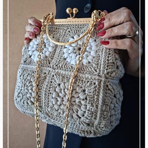 Puede incluir: Un bolso de mano de ganchillo beige con una cadena y un cierre dorados. El bolso presenta un patrón floral con detalles en blanco. El cierre tiene un diseño decorativo. El bolso es sostenido por una persona.