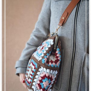 Puede incluir: Bolso tejido a ganchillo en marrón, azul, rosa y blanco, con diseño de cuadrados de la abuela. Tiene una correa de cuero marrón y un cierre metálico. El bolso lo lleva una persona con un abrigo gris. Dimensiones: aprox. 25 cm x 25 cm.