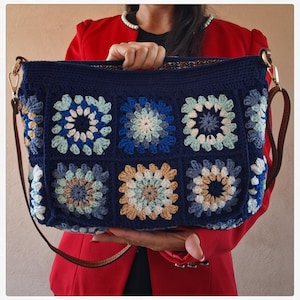 May include: A rectangular crochet shoulder bag featuring a dark navy blue base with a grid of six square motifs. Each motif displays a circular pattern in shades of light blue, white, and tan. The bag includes a brown leather-like shoulder strap.