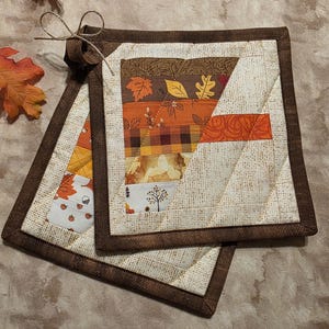 May include: Two square quilted pot holders featuring a patchwork design of autumn-themed fabrics in shades of orange, brown, yellow, and cream. The pieces are bordered by a dark brown fabric and include a small loop with a tied string for hanging.