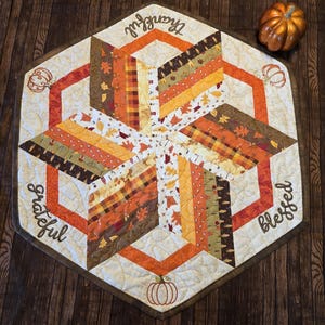 May include: A quilted, hexagonal table runner with a fall theme. The runner features a starburst design with orange, brown, and yellow fabrics. The words "Thankful", "Grateful", and "Blessed" are embroidered on the edges. A small pumpkin is also present.