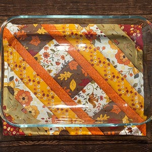 May include: A rectangular clear glass baking dish sits atop a fabric hot pad featuring a diagonal patchwork design. The fabric displays an autumn-themed pattern with orange, yellow, and brown colors, including illustrations of fall leaves and flowers.