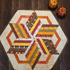 May include: A hexagonal quilted table runner featuring a star pattern made of fabric strips in shades of orange, yellow, brown, and cream. The design includes autumn leaf prints, plaid, and floral motifs with a cream-colored background.