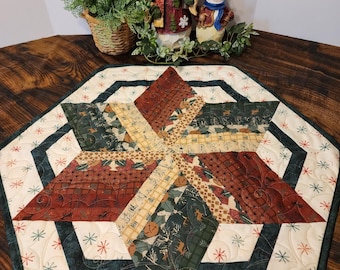 May include: A hexagonal table runner with a starburst design in shades of green, brown, and cream. The runner is quilted with a snowflake pattern and has a dark green border.